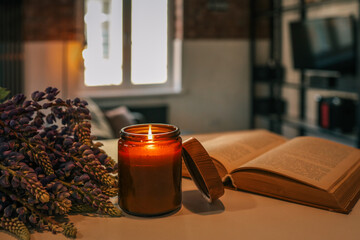 Lit candle, purple flowers, and an open vintage book on a light wooden table in a cozy loft-style room. Warm and peaceful atmosphere with natural light and rustic brick walls