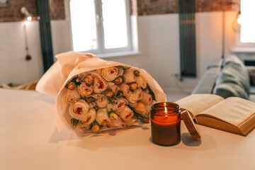 A bouquet of pastel pink roses wrapped in paper, placed beside a burning candle and an open book on...