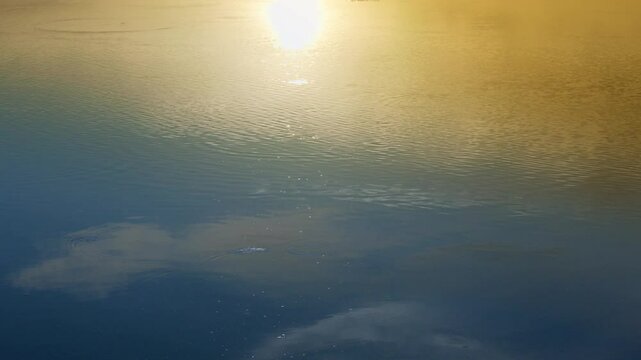 Viewed from above, golden sunlight reflects across tranquil waters beneath a shifting cloudscape&mdash;blending deep blue tones with warm hues, the scene evokes a quiet moment of natural beauty and atmosphe