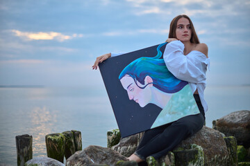 Young woman stands on rocky pier holding portrait she painted, set against calm sea. Painting...