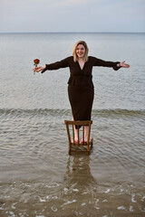 Young woman in black outfit stands confidently on chair in ocean, holding rose. Scene evokes themes...