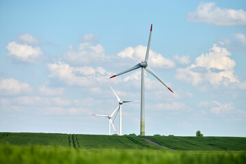 Dirt road leads through green field with two wind turbines towering under blue sky filled with...