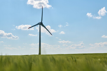 Wind turbine stands tall in lush green wheat field beneath bright blue sky with fluffy white clouds...
