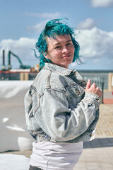 Young woman with teal hair and denim jacket stands near seaside area. Bright daylight illuminates...