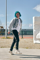 Young woman with blue hair and denim jacket walking happily near urban artwork. Bright sunlight....