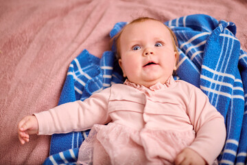 Baby girl with light brown hair, caucasian ethnicity, wearing pink dress, lies on blue blanket with...