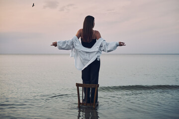 Young woman stands on chair in calm ocean waters at dusk, arms outstretched. Tranquil scene...