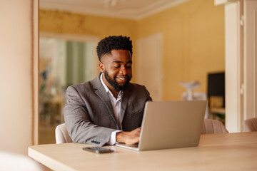 Portrait of young businessman smiling and working online over laptop at desk in corporate office