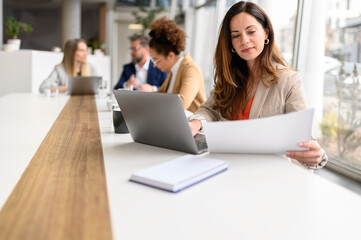 Focused female lawyer reading document and working over laptop in board room with colleagues in the...