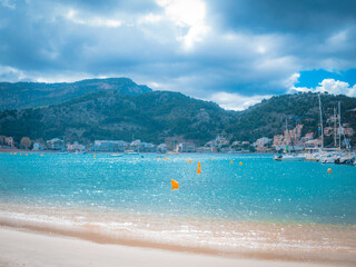 Port de Sóller in Mallorca, Spain, features turquoise waters, a sandy beach, and mountain views—an idyllic Mediterranean harbor under a partly cloudy sky perfect for travel imagery.