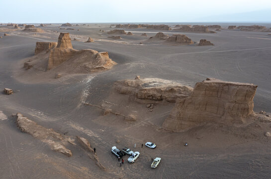 Aerial view of sun-baked yardangs casting long shadows across the arid landscape, a small group gathered near vehicles, their presence dwarfed by the immensity, Kerman, Kerman Province, Iran.