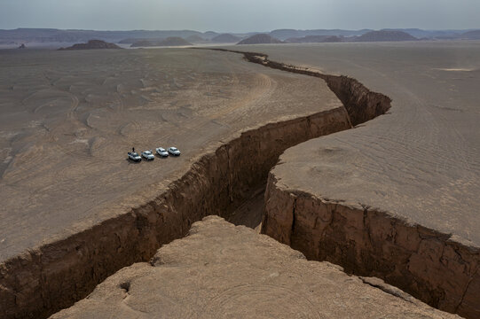 Aerial view of a dramatic canyon slicing through the parched earth, vehicles dwarfed beside the colossal fissure, a testament to geological forces, Kerman, Kerman Province, Iran.