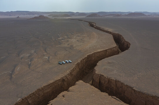 Aerial view of a profound chasm slices through the earth, juxtaposed against a fleet of vehicles, echoing the vastness of the desert, Kerman, Kerman Province, Iran.