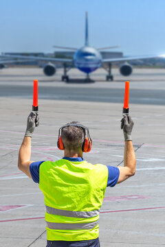 Rear view of aircraft marshaller in PPE guiding airplane on runway with marshalling wands at airport.