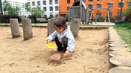 Little girl playing with sand in a modern urban playground, wearing a light jacket and using toy shovel and bucket, concept of childhood and outdoor activity