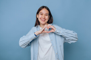 joyful young woman forms a heart shape with her hands, celebrating love and positivity on light...