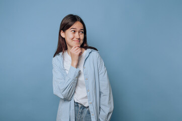 young woman playfully touches her chin with a smile, embodying a mischievous secret on light blue...