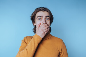 young man covers his mouth in surprise, conveying astonishment and shock while posing on blue...