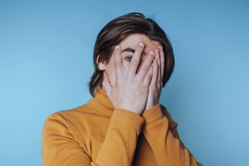 young man in a turtleneck hides his face in his hands, showcasing shyness and vulnerability on blue...
