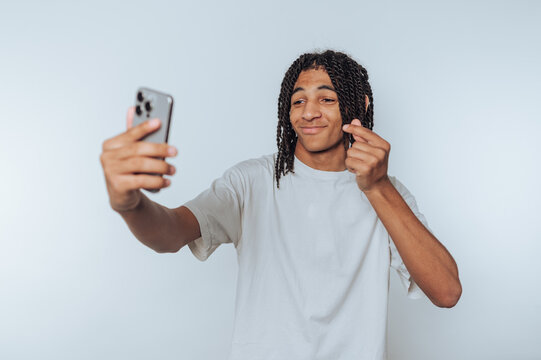 young man joyfully celebrates while holding a smartphone while making video call and showing heart gesture on white background