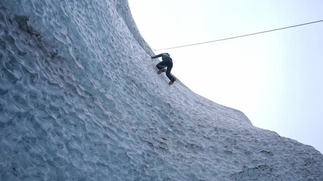 A climber, equipped with an ice axe and rope, scales a majestic, textured blue ice wall of a glacier, showcasing the extreme adventure amidst the vast ice formations of Vatnaj&ouml;kull, Iceland.