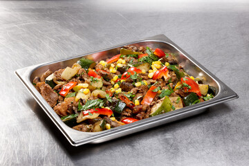 A mixture of grilled meat and fresh vegetables including zucchini, bell peppers, corn, and herbs is displayed in a metal serving tray. The vibrant colors create an appetizing presentation for diners