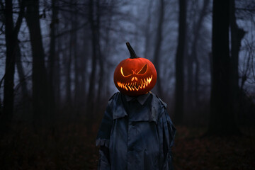 Smiling figure in an old torn cloak with a glowing from inside pumpkin jack-o-lantern head in dark...