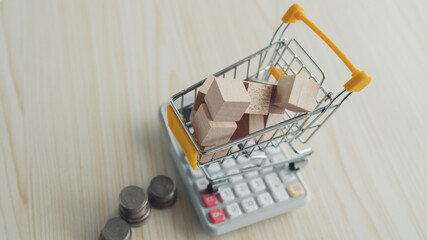 Mini shopping cart filled with wooden blocks sits on a calculator, next to stacks of coins,...