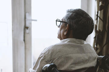 Close picture of indian disabled senior old man sits in the wheelchair looking outside on balcony while thinking something after retirement at home
