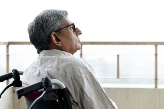 Close picture of indian disabled senior old man sits in the wheelchair looking outside on balcony while thinking something after retirement at home