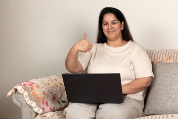 Indian plus size overweight female businesswoman sit relaxing on sofa smiling hold hand show thumb up using laptop work from home