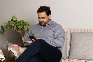 Fototapeta premium Indian handsome smiling man taking on phone while relaxing using laptop computer working and video conference meeting at work from home