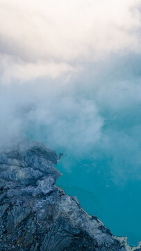 Aerial view of the rugged, dark volcanic rocks meet the vibrant turquoise crater lake under a blanket of ethereal white mist, Ijen Volcano Crater, East Java, Indonesia.