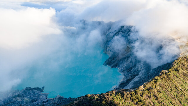 Aerial view of the mesmerizing turquoise crater lake nestled amidst rugged, volcanic terrain, shrouded in ethereal clouds, Ijen Volcano Crater, East Java, Indonesia.