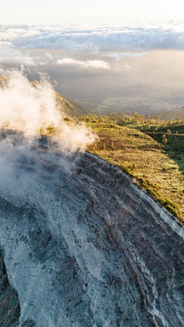 Aerial view of a rugged mountain face where mist delicately veils the slopes, blending with the soft glow of the morning light, Ijen Volcano Crater, East Java, Indonesia.