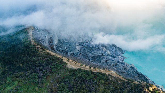 Aerial view of clouds clinging to the volcanic landscape, a path winding along the rim, and turquoise waters, Ijen Volcano Crater, East Java, Indonesia.