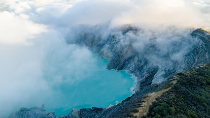 Aerial view of the turquoise crater lake nestled amidst the rugged terrain of Ijen Volcano, shrouded in ethereal clouds, Ijen Volcano Crater, East Java, Indonesia.
