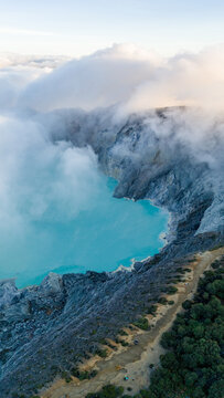 Aerial view of the stunning turquoise lake nestled within the rugged Ijen Volcano Crater, shrouded in ethereal clouds and mist, Ijen Volcano Crater, East Java, Indonesia.