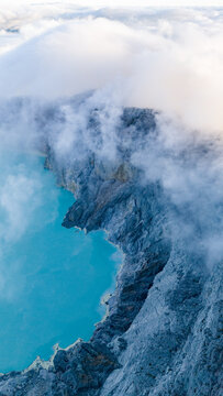 Aerial view of the vibrant turquoise crater lake nestled amidst jagged, rocky terrain, shrouded in ethereal mist and clouds, Ijen Volcano Crater, East Java, Indonesia.
