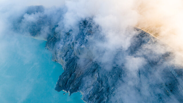 Aerial view of ethereal clouds swirling around the rugged, volcanic landscape of Ijen Crater, contrasting starkly with the turquoise lake below, Ijen Volcano Crater, East Java, Indonesia.