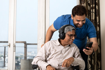 Indian son with mobile phone looking at senior dad smiling while listening music in headphones at...