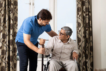 Indian caring con taking care of senior father in wheelchair in after retirement. Asian happy disabled man gesturing while talking to smiling indian son in living room at home
