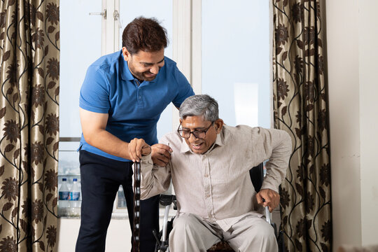 Indian caring con taking care of senior father in wheelchair in after retirement. Asian happy disabled man gesturing while talking to smiling indian son in living room at home