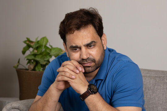 Indian unhappy sad man sitting on sofa alone at home, suffering from depression, loneliness or anxiety, copy space. Permacrisis, midlife crisis, financial troubles, lack of money and health problems