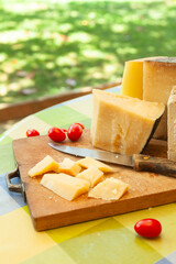Chunky pieces of hard cheese with knife and cherry tomatoes on wooden board in bright outdoor...