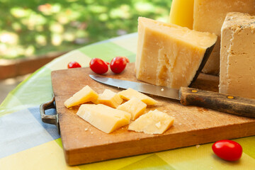 Chunky pieces of hard cheese with knife and cherry tomatoes on wooden board in bright outdoor...