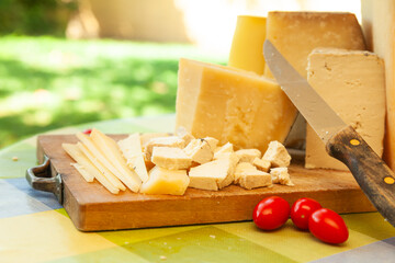 Assorted blocks and slices of hard cheese on wooden board with knife and tomatoes in outdoor summer...