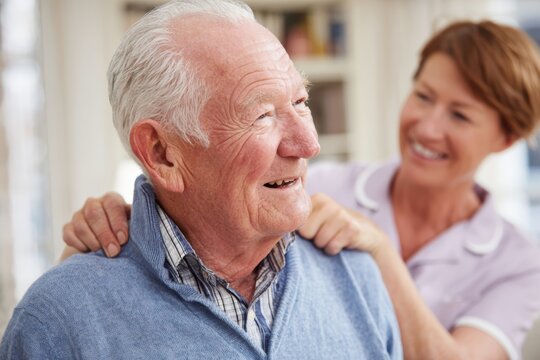 Smiling elderly man receiving a caring shoulder massage from a friendly female nurse in a cozy home setting.
