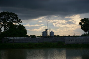 Workers Installing Equipment on Industrial Silos under Dramatic Cloudy Sky near a Rural Pond
