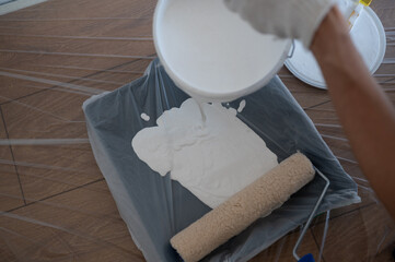 Worker pouring white paint into a paint tray covered with a plastic sheet, preparing to paint a wooden floor with a paint roller